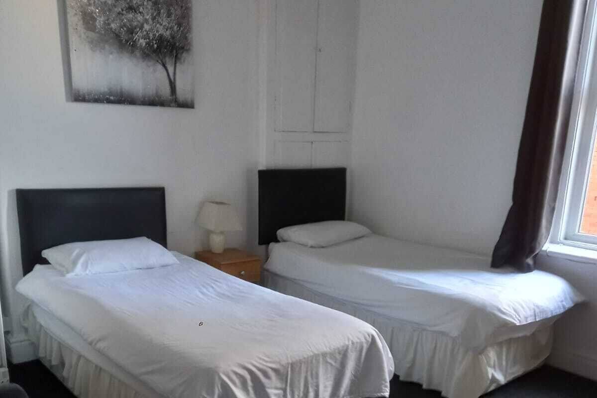 A hotel room with two single beds, a nightstand with a lamp, a painting on the wall, and a window with curtains.