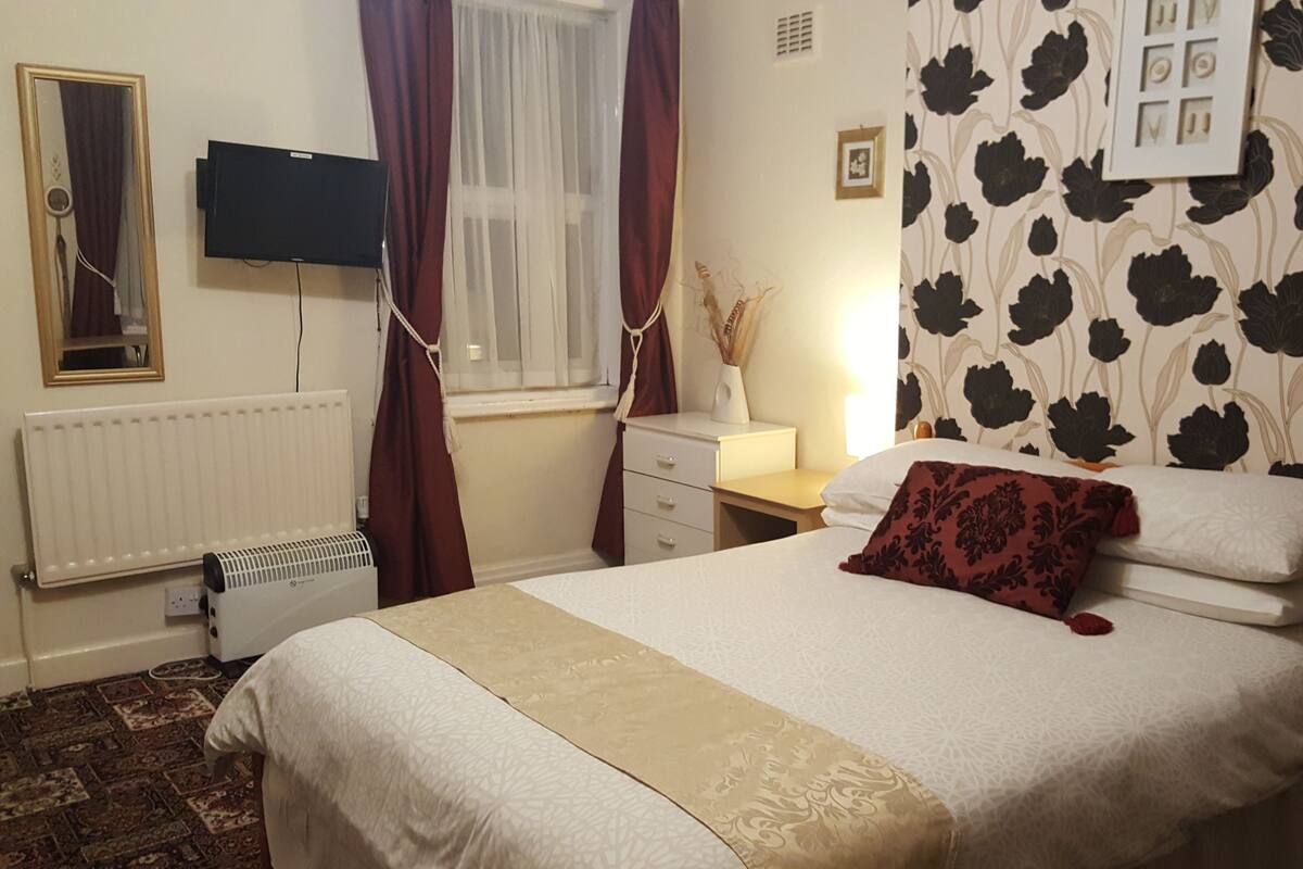 A neatly arranged bedroom with a bed, a bedside table, a mirror, a radiator, and a TV mounted on the wall.