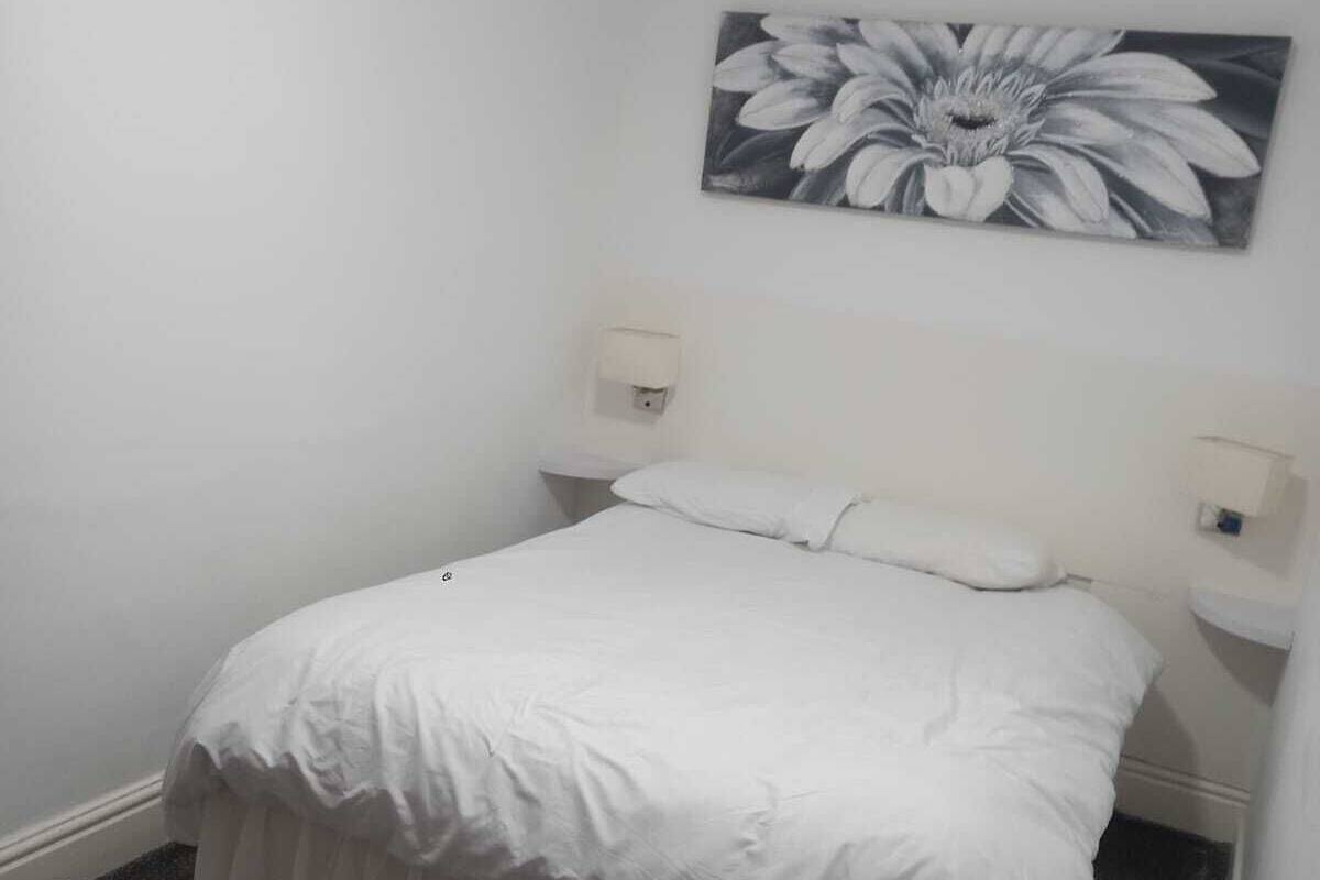 A bedroom with a single bed, white bedding, a gray carpet, and a wall-mounted painting of a flower.
