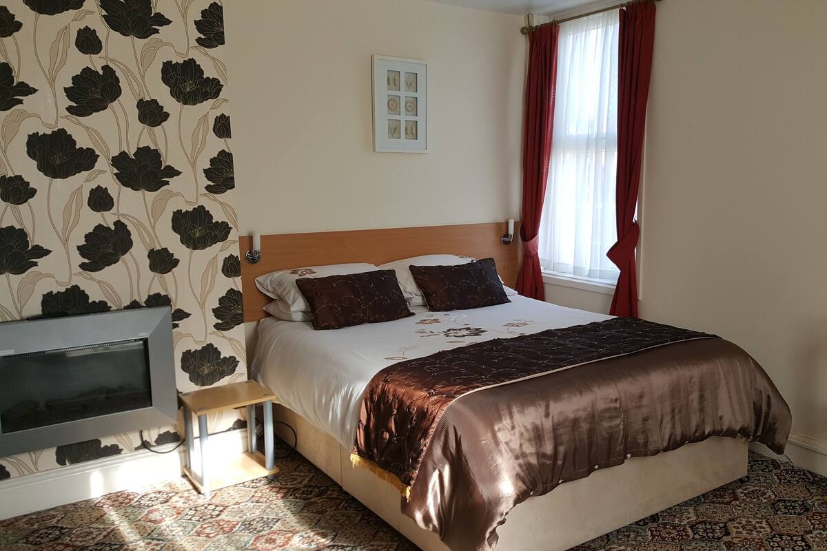 A bedroom with a bed, a TV, a window with red curtains, and a wall with a floral pattern.