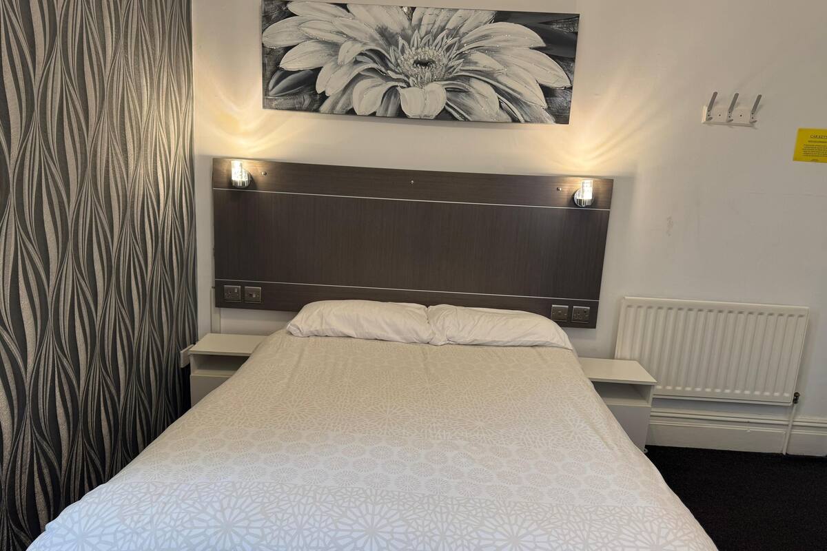 A neatly made bed with a patterned bedspread, a headboard with wall-mounted lights, a radiator, and a black and white floral artwork above the bed.