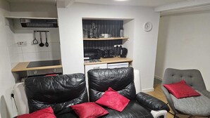 Een ruimtelijke zithoek met een kitchenette, een zwarte leren bank met rode kussens, een grijze fauteuil en een kleine eettafel.