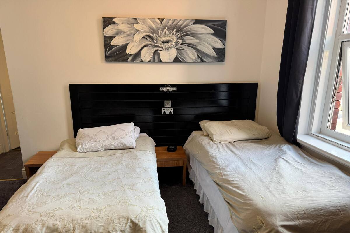 A bedroom with two single beds, a black headboard, a wall-mounted flower painting, and a window with curtains.