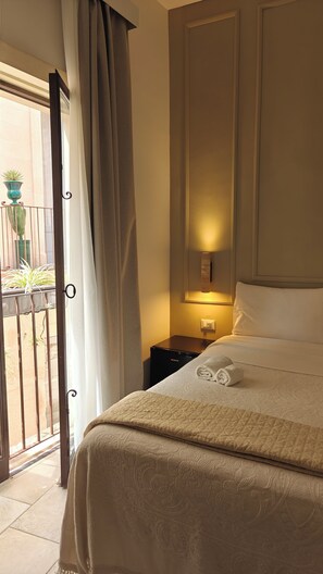 A hotel room with a bed, a nightstand, a wall-mounted lamp, and a view through a curtain to an outdoor area with plants.