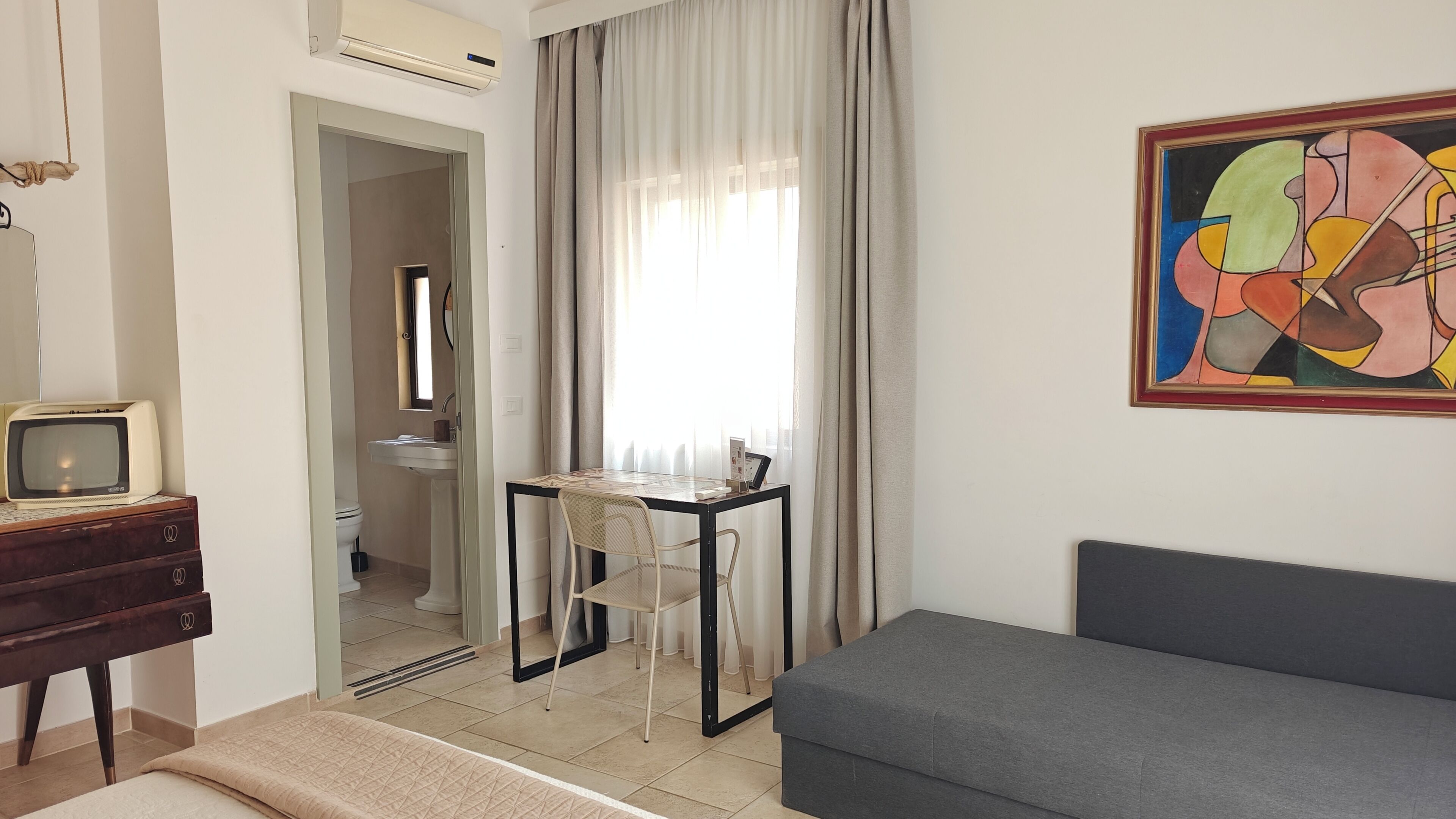 A room with a bed, a desk with a chair, a television, a painting, and a bathroom visible through an open door.
