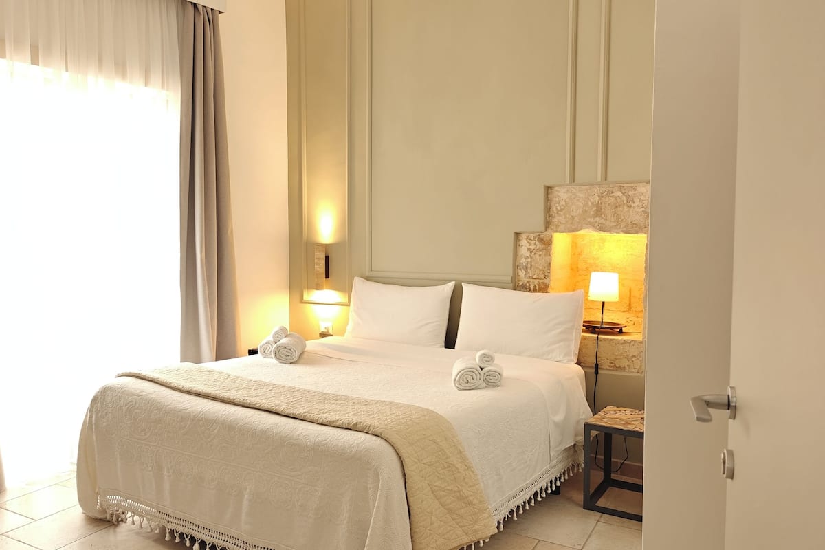 A hotel room with a bed, two bedside lamps, a nightstand, and a view of the outside through a window with curtains.