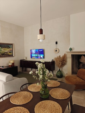 A living room with a TV, sofa, dining table with placemats, a vase with flowers, and a fireplace.