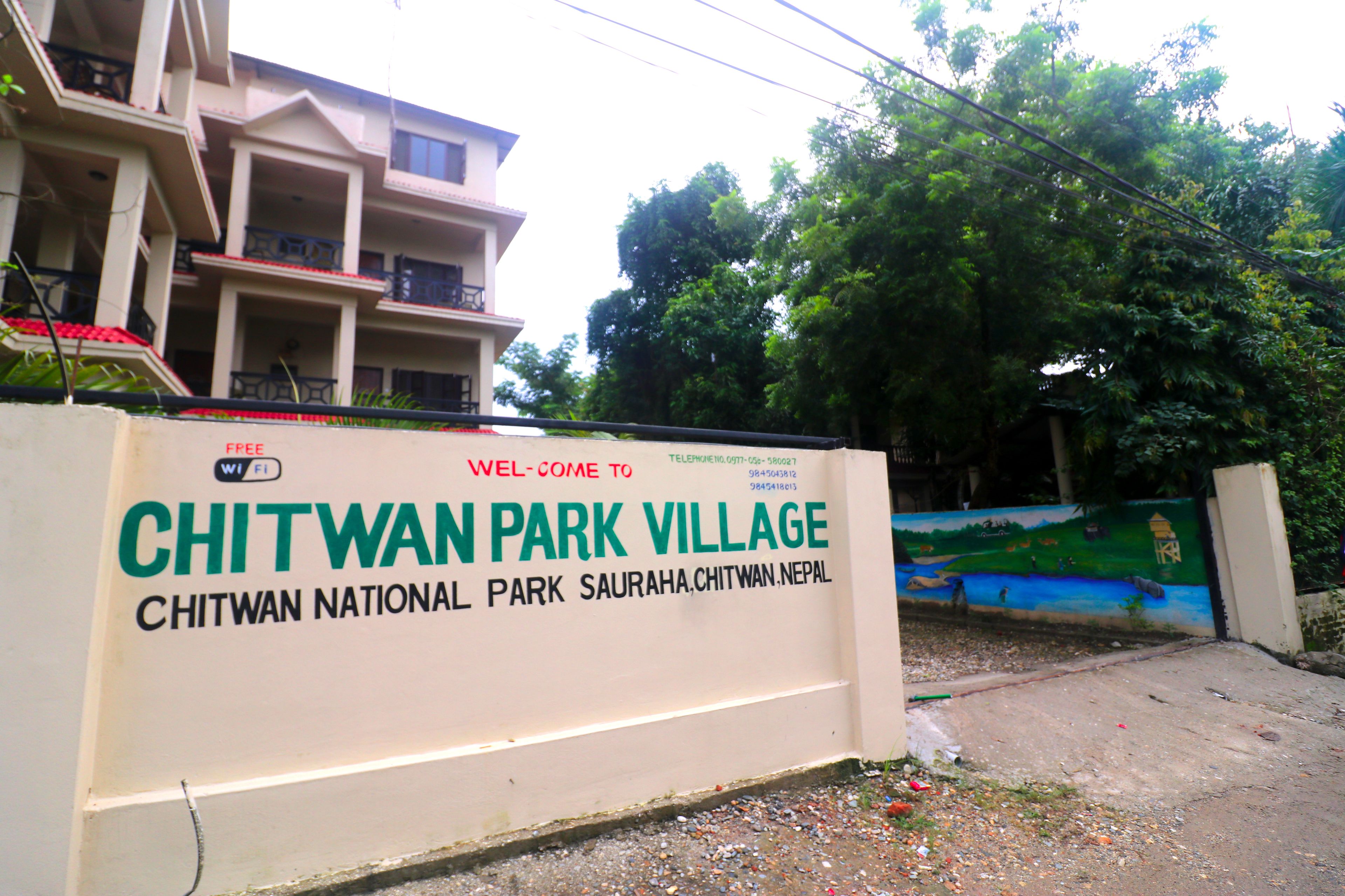A sign for Chitwan National Park Village with a view of a building and trees in the background.
