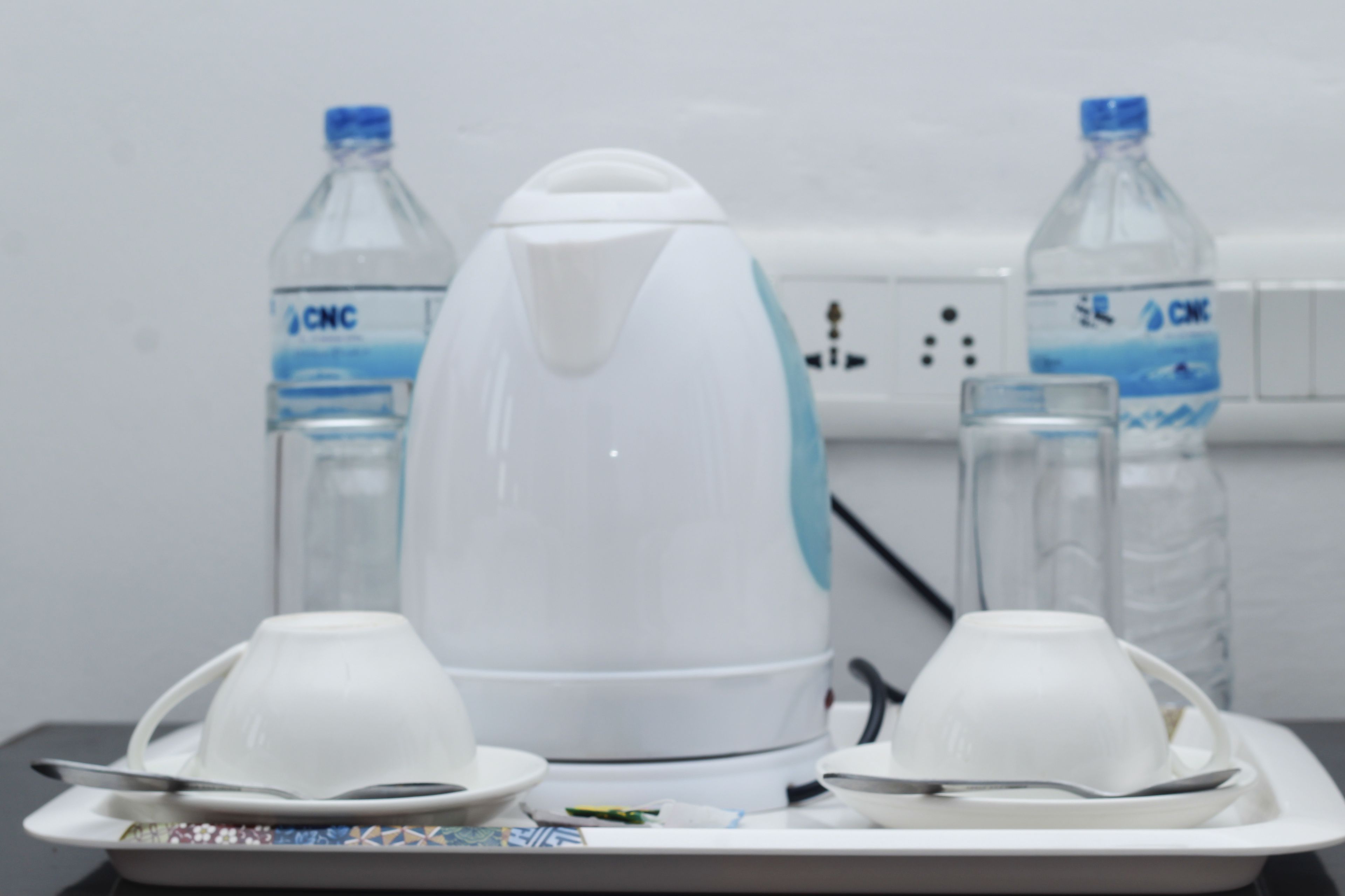 A kitchen area with a white electric kettle, two water bottles labeled 'CNC', and a tray with two white cups and a spoon.