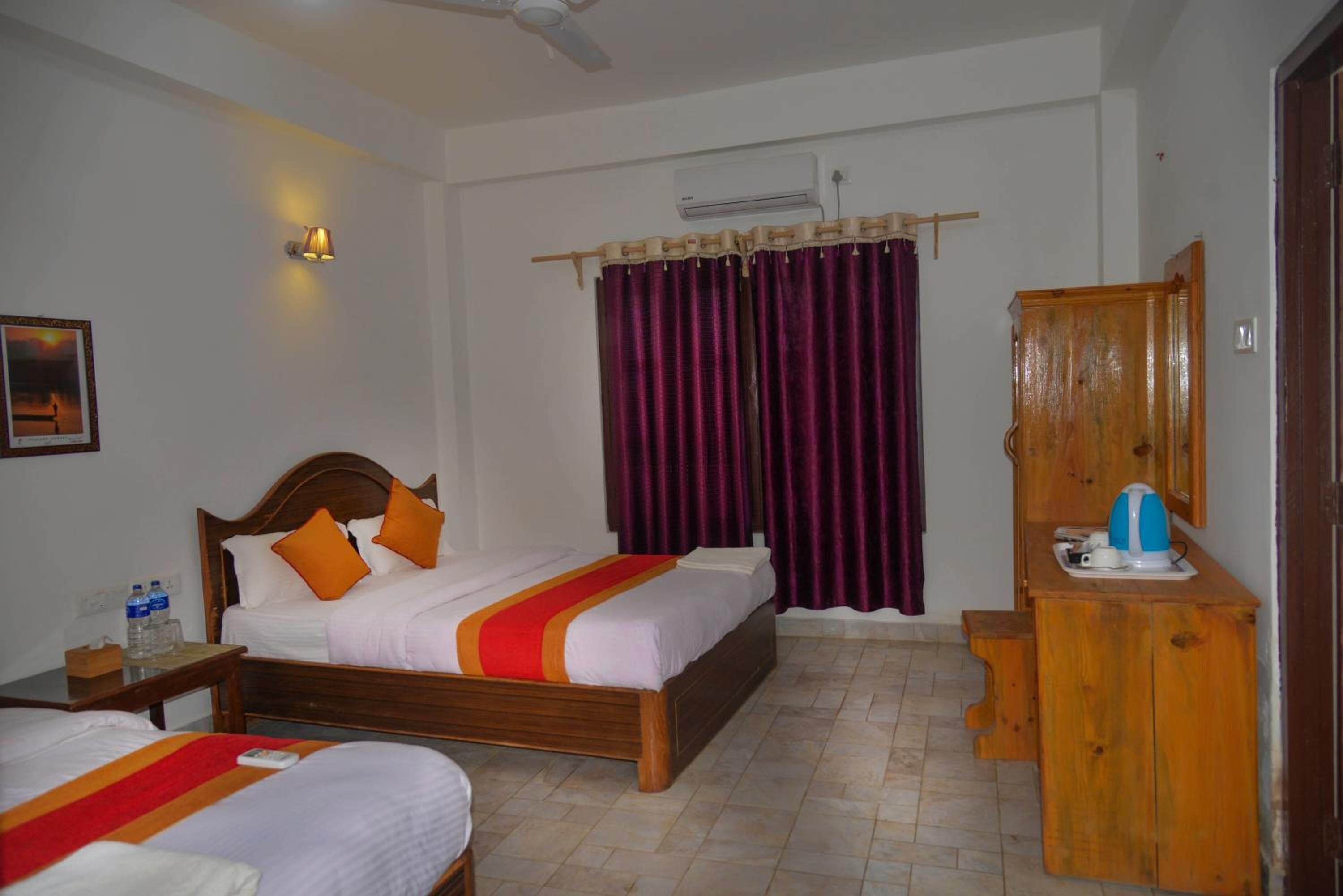 A hotel room with two beds, a wooden wardrobe, a bedside table with a lamp, and an air conditioning unit.