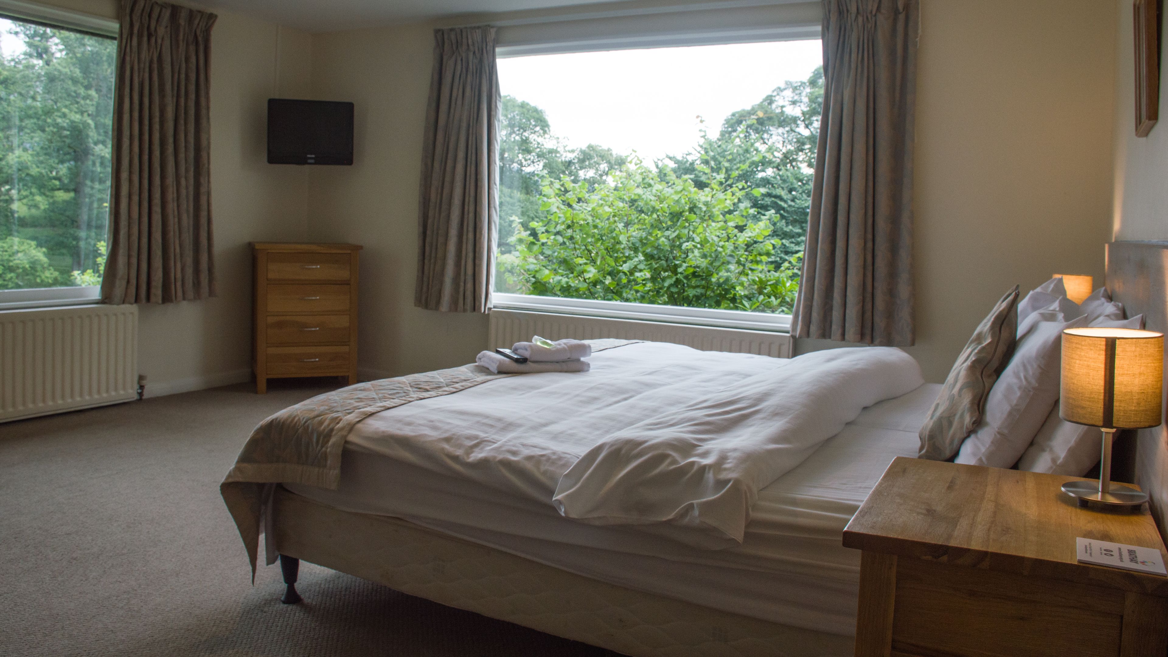 A neatly made bed with a blanket and pillows, a bedside table with a lamp, a wooden dresser, and a television on the wall.