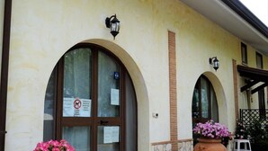 Ein Gebäude mit zwei großen Bogenöffnungen, jeweils mit einem Schild darüber. Vor den Türen befinden sich zwei große Terrakottatöpfe mit rosa Blumen.