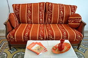 A red sofa with floral patterns and a matching pillow, a white table with a red vase and a magazine, and a patterned floor. - Colonna Apartment with Terrace (Florence)