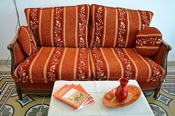 Un canapé rouge orné de motifs floraux, accompagné d’un coussin assorti, une table blanche sur laquelle reposent un vase rouge et un magazine, et un sol à motifs.