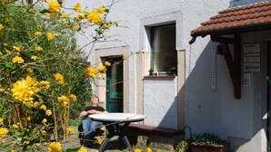 A small outdoor seating area with a table and chairs, a white building with a tiled roof, and a garden with yellow flowers. - Grüne Idylle im Dresdener Süden (Bannewitz)