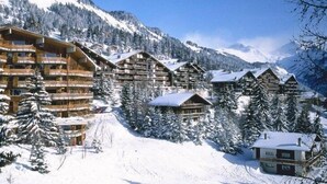Un chalet de montagne enneigé, composé de plusieurs bâtiments en bois, entouré d’arbres et de montagnes couverts de neige.
