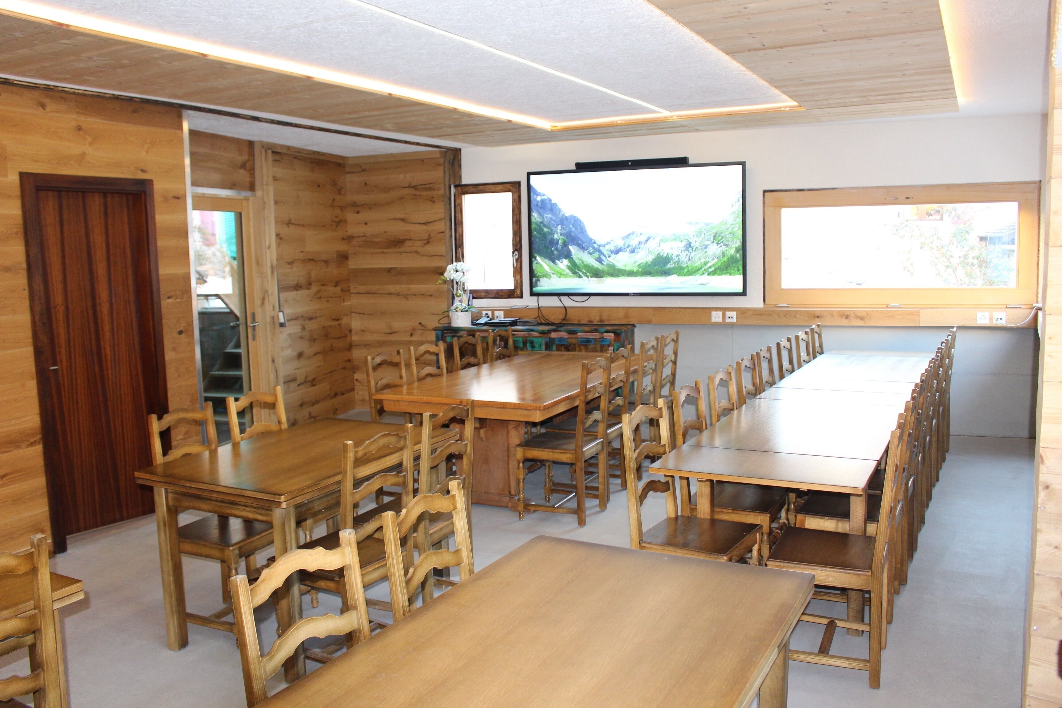 A long wooden dining table with chairs, a television mounted on the wall, and a window with a mountain view.