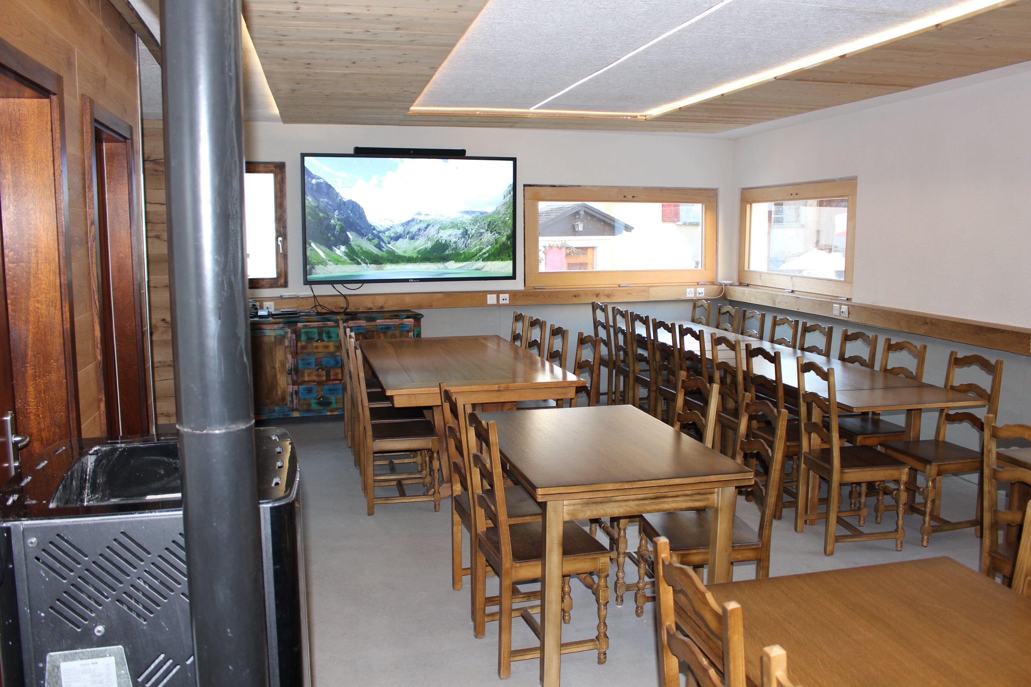 Un espace repas avec des tables et des chaises en bois, un téléviseur fixé au mur et une grande fenêtre donnant sur l’extérieur.
