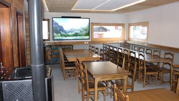 Un espace repas avec des tables et des chaises en bois, un téléviseur fixé au mur et une grande fenêtre donnant sur l’extérieur.