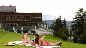 Un centre de bien-être et de soins avec un bâtiment moderne, une piscine et un espace verdoyant où deux personnes se détendent sur une serviette.
