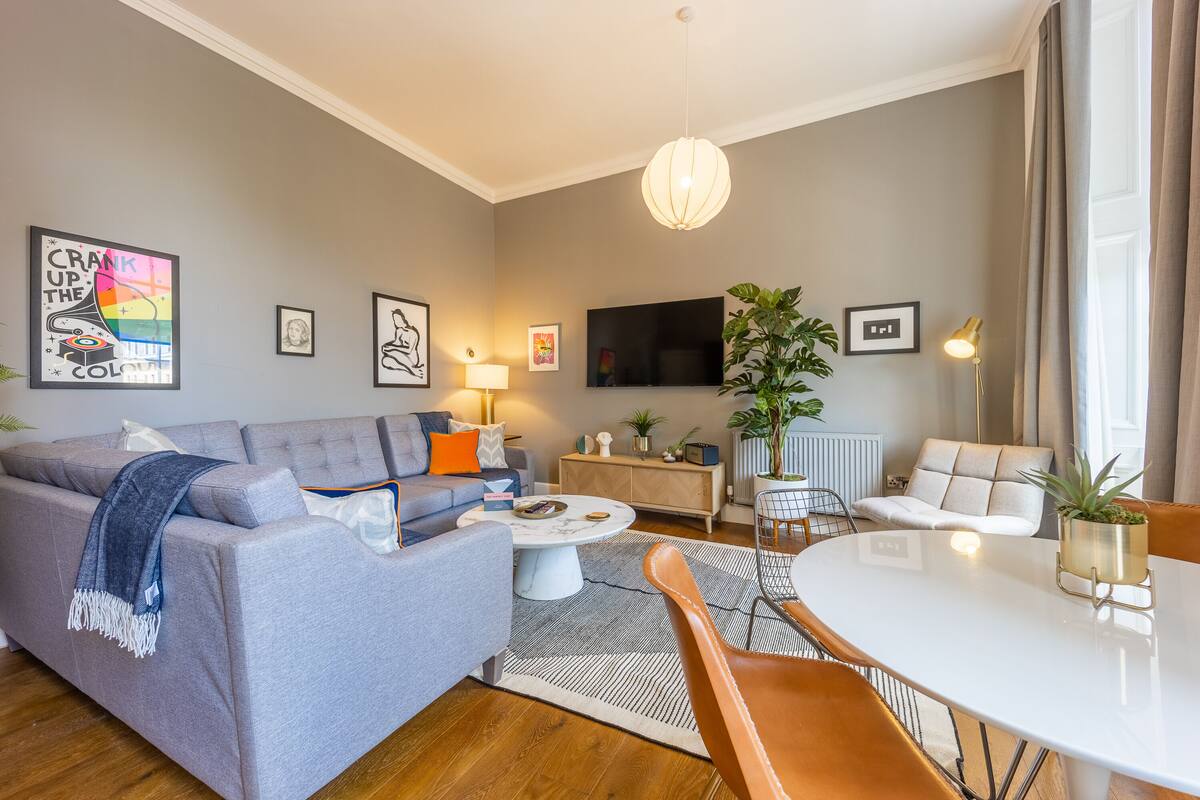A modern living room with a grey sofa, a coffee table, and a dining table with chairs. There are framed pictures on the walls, a TV, and a potted plant.
