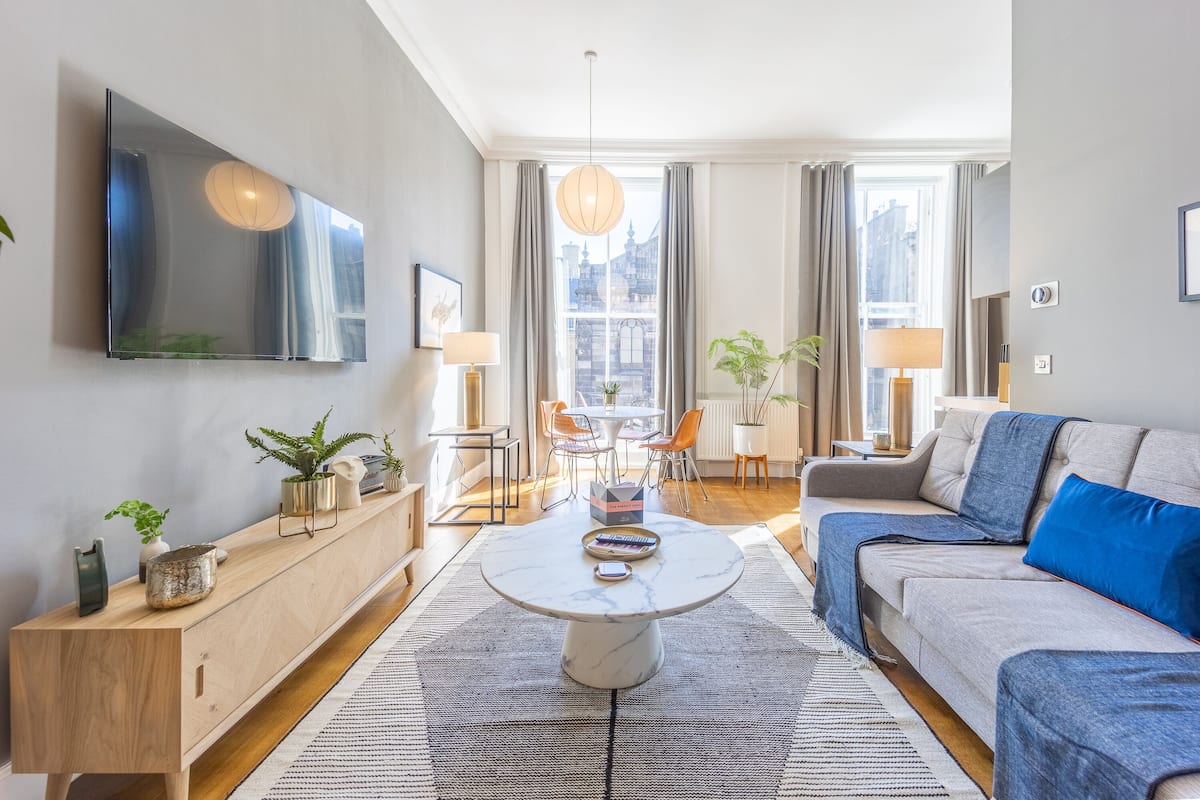 A modern living room with a sofa, coffee table, and a TV mounted on the wall. There is a dining area with a table and chairs, and a view of buildings outside the window.