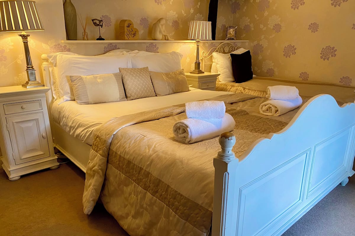 Un lit bien fait, recouvert d’une courtepointe matelassée, deux tables de chevet avec des lampes, et un mur orné d’un papier peint à motifs floraux.
