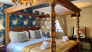 A four-poster bed with a canopy, a wooden headboard, floral wallpaper, and a bedside table with a lamp.