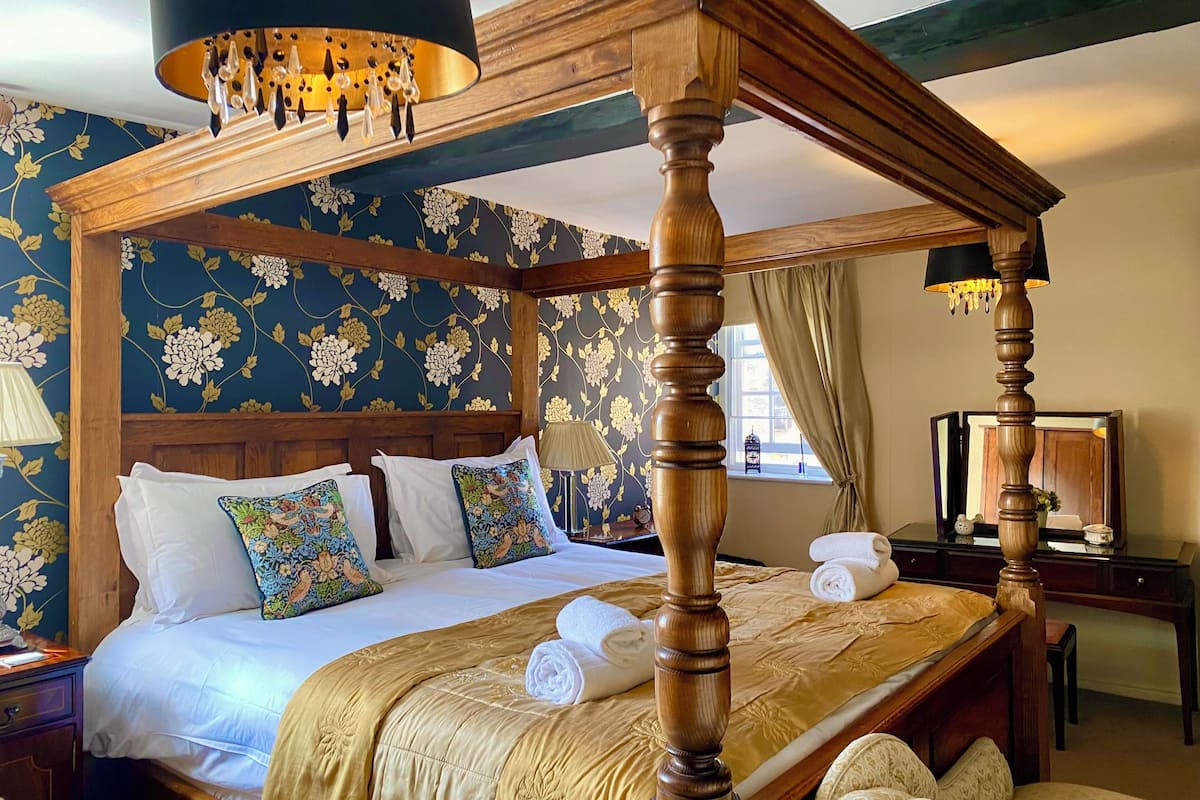 Un lit à baldaquin avec des poteaux aux quatre coins, une tête de lit en bois, un papier peint à motifs floraux et une table de chevet avec une lampe.