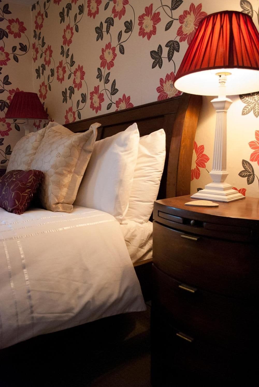 A neatly made bed with white bedding and several pillows, a wooden headboard, a bedside table with a lamp, and a wall with floral wallpaper.