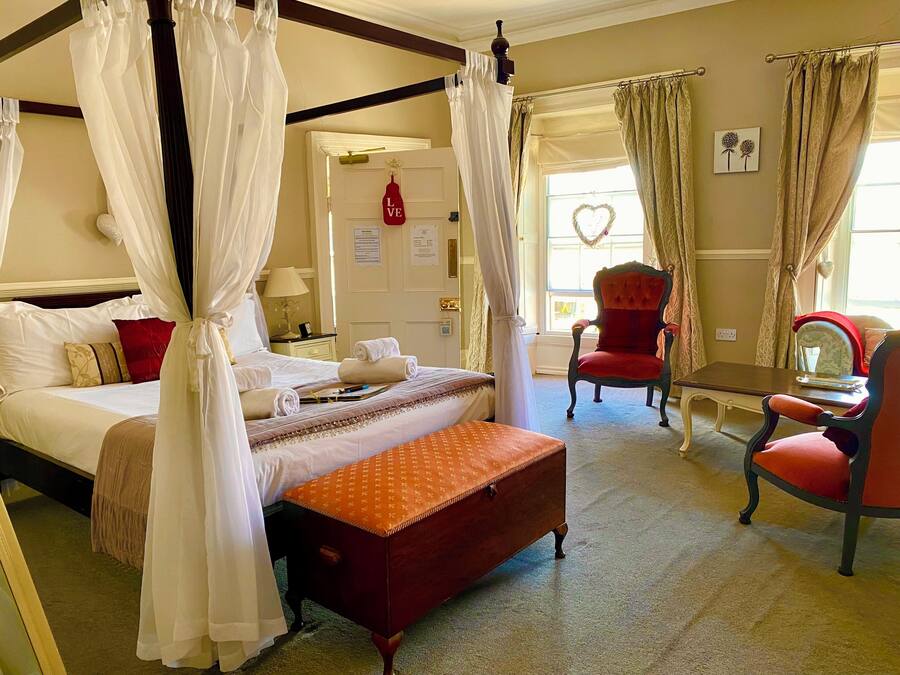 Una cama con dosel, un sillón rojo, una mesita y un adorno con forma de corazón en la ventana.