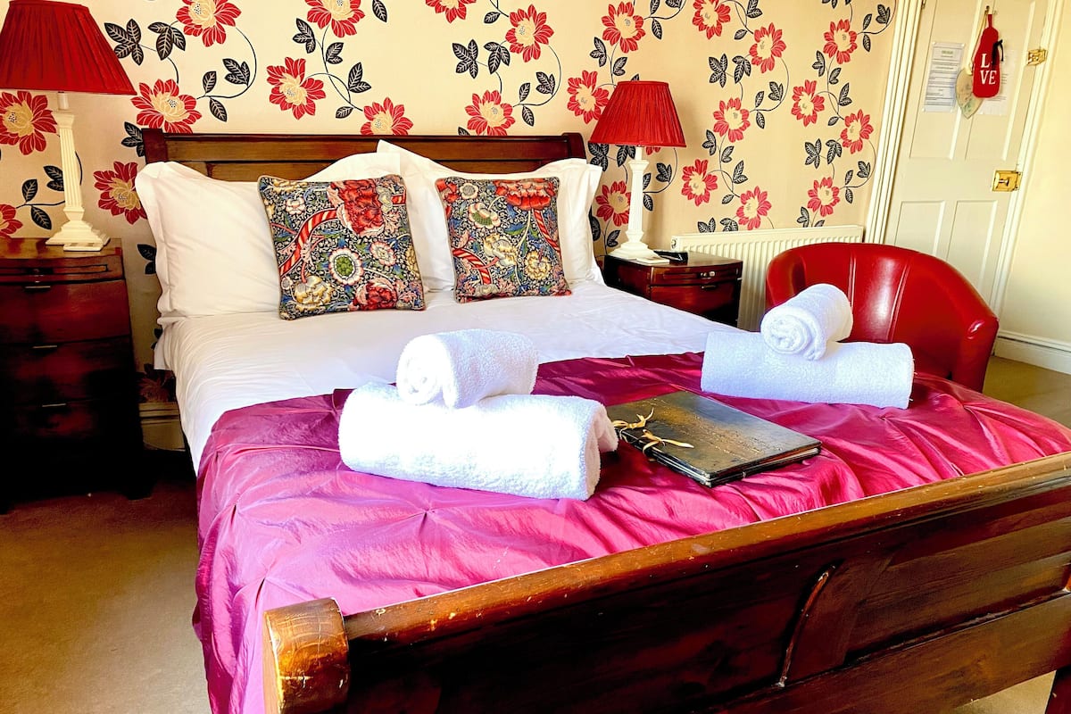 Un lit bien fait, recouvert d’une courtepointe rouge, avec deux oreillers à motifs floraux et deux serviettes blanches.