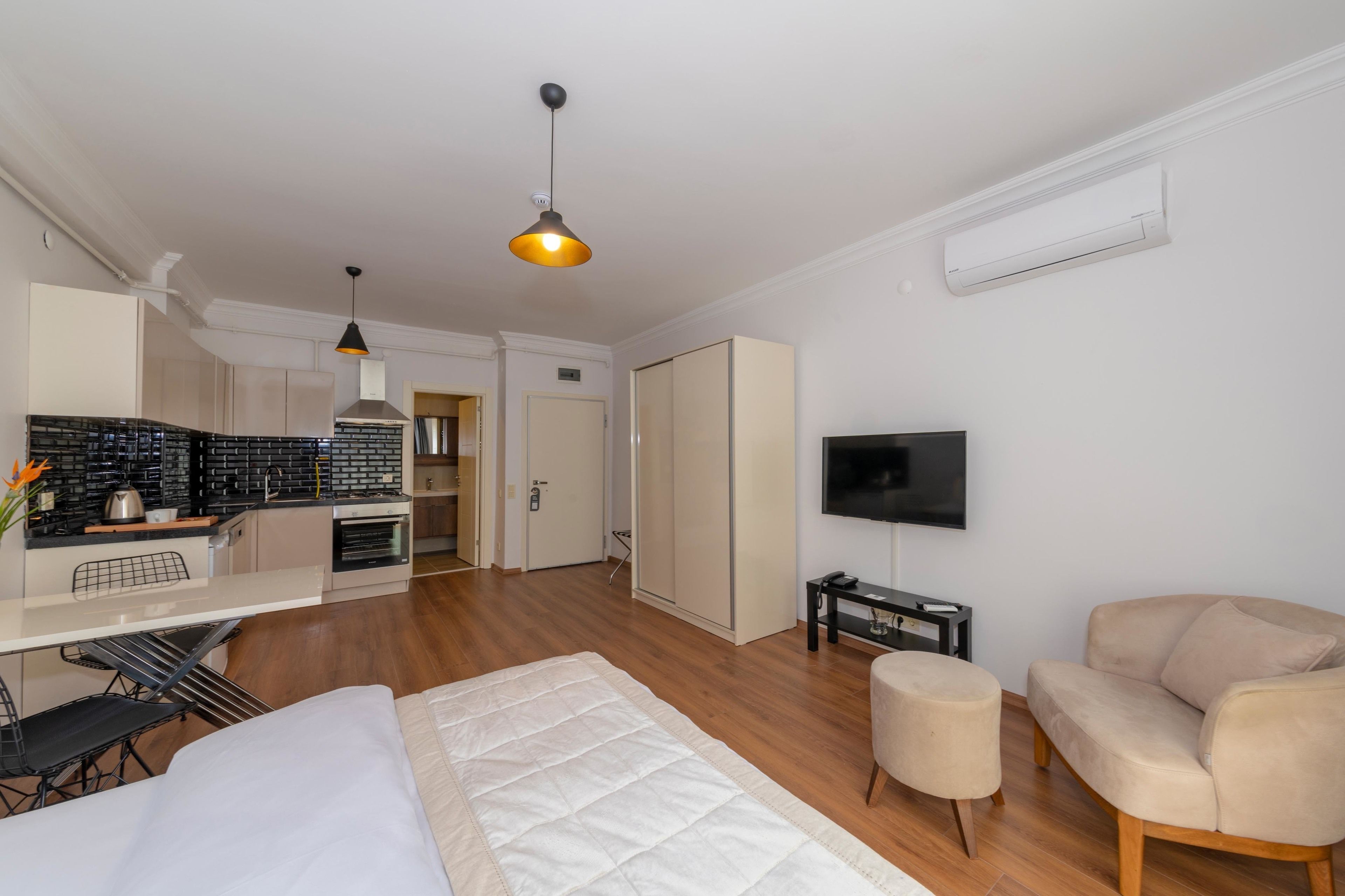 A modern, well-lit room with a kitchenette, a flat-screen TV, a sofa, and a bed.
