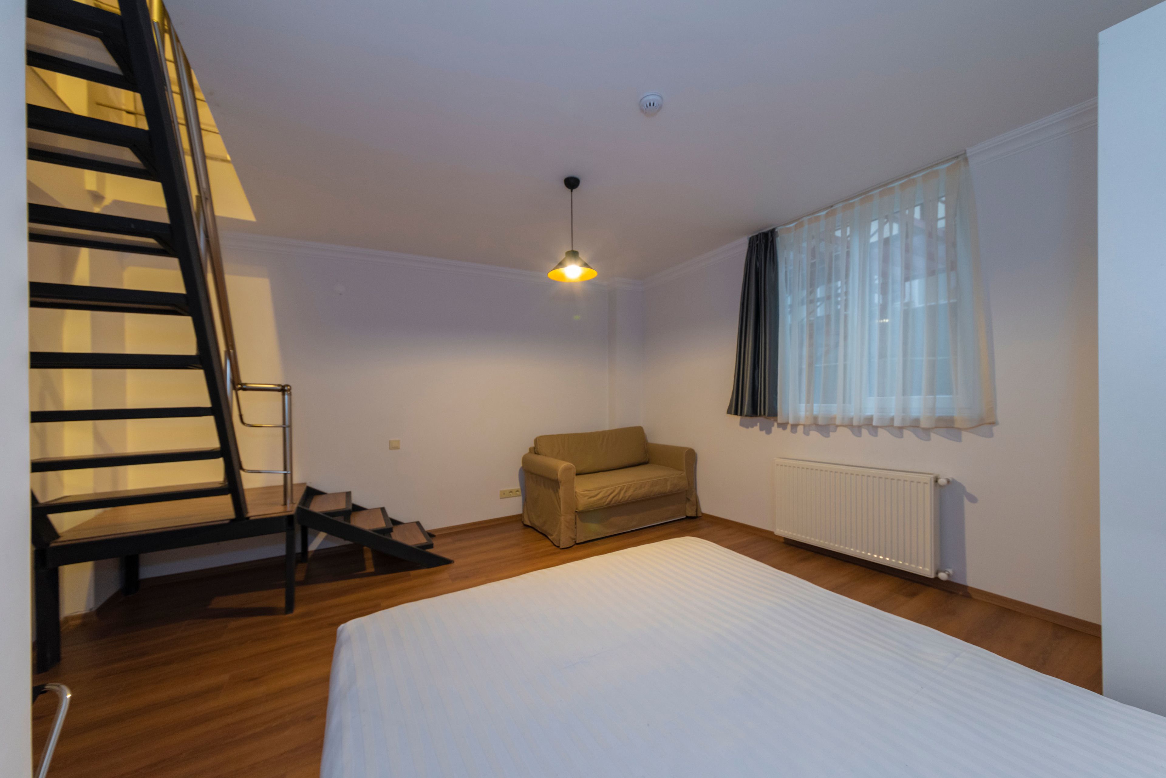 A bedroom with a bed, a chair, a radiator, a window with curtains, and a staircase.