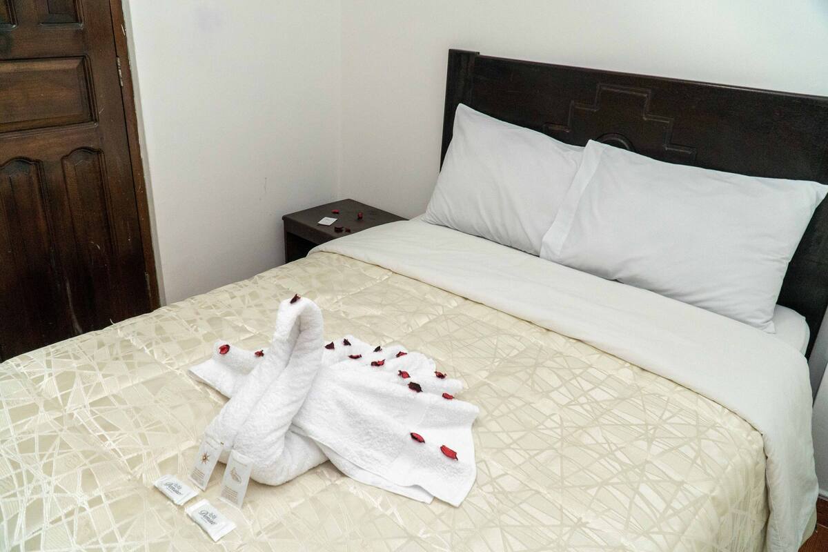 Cama bem arrumada com colcha bege estampada, almofadas brancas e uma toalha branca dobrada com pétalas de flores vermelhas sobre uma mesa de cabeceira de madeira.