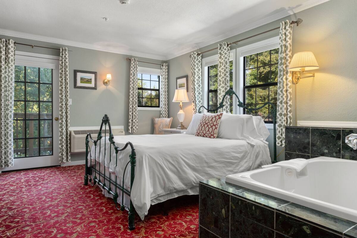 A bedroom with a large bed, a separate bathroom with a bathtub, and a view of trees through the windows.