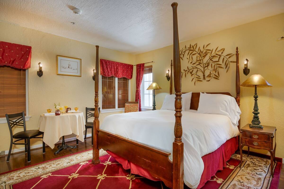 A four-poster bed with a red and white bedspread, a wooden nightstand with a lamp, a small table with a chair, and a decorative wall piece.
