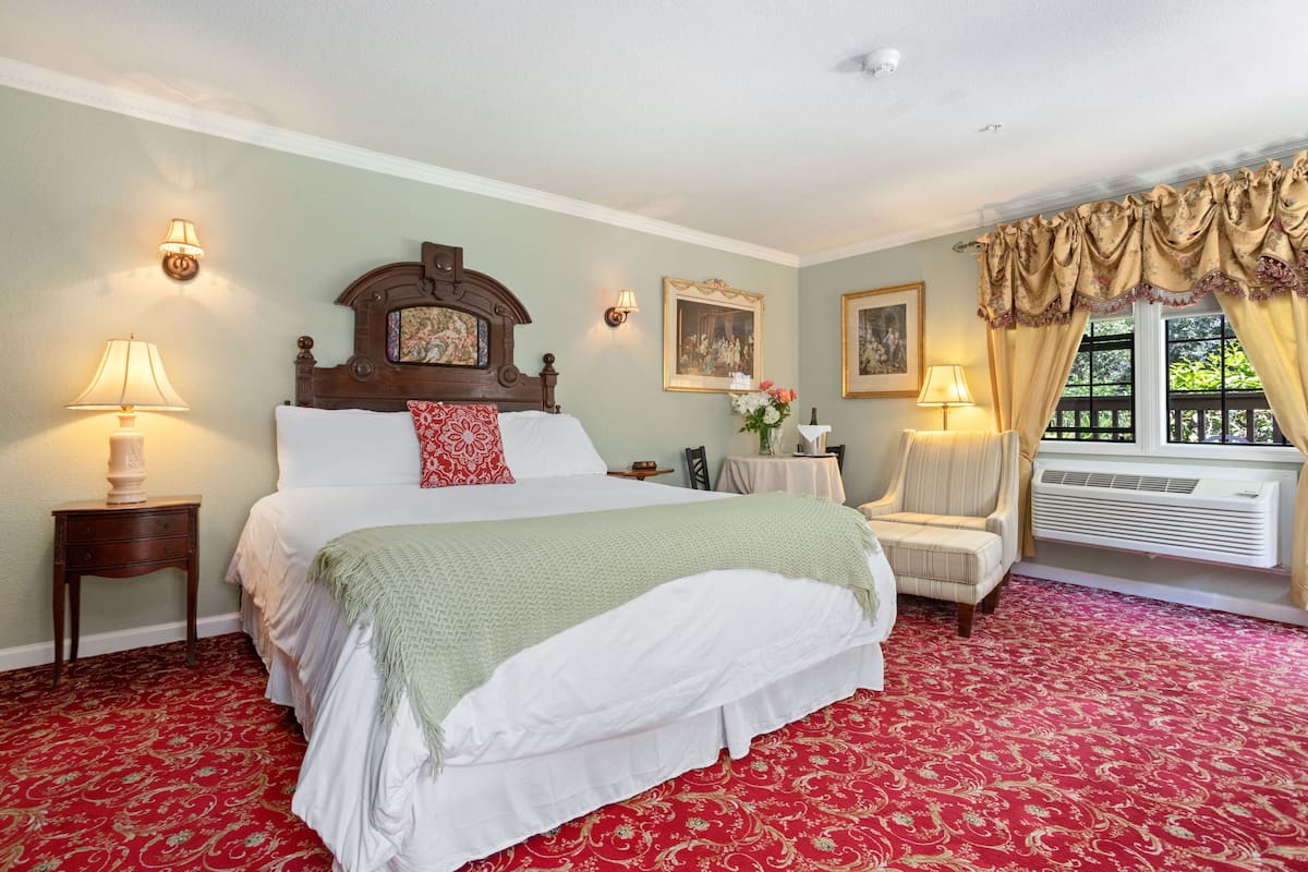 A hotel room with a large bed, two bedside tables with lamps, a chair, a small table with a vase, and a window with curtains.