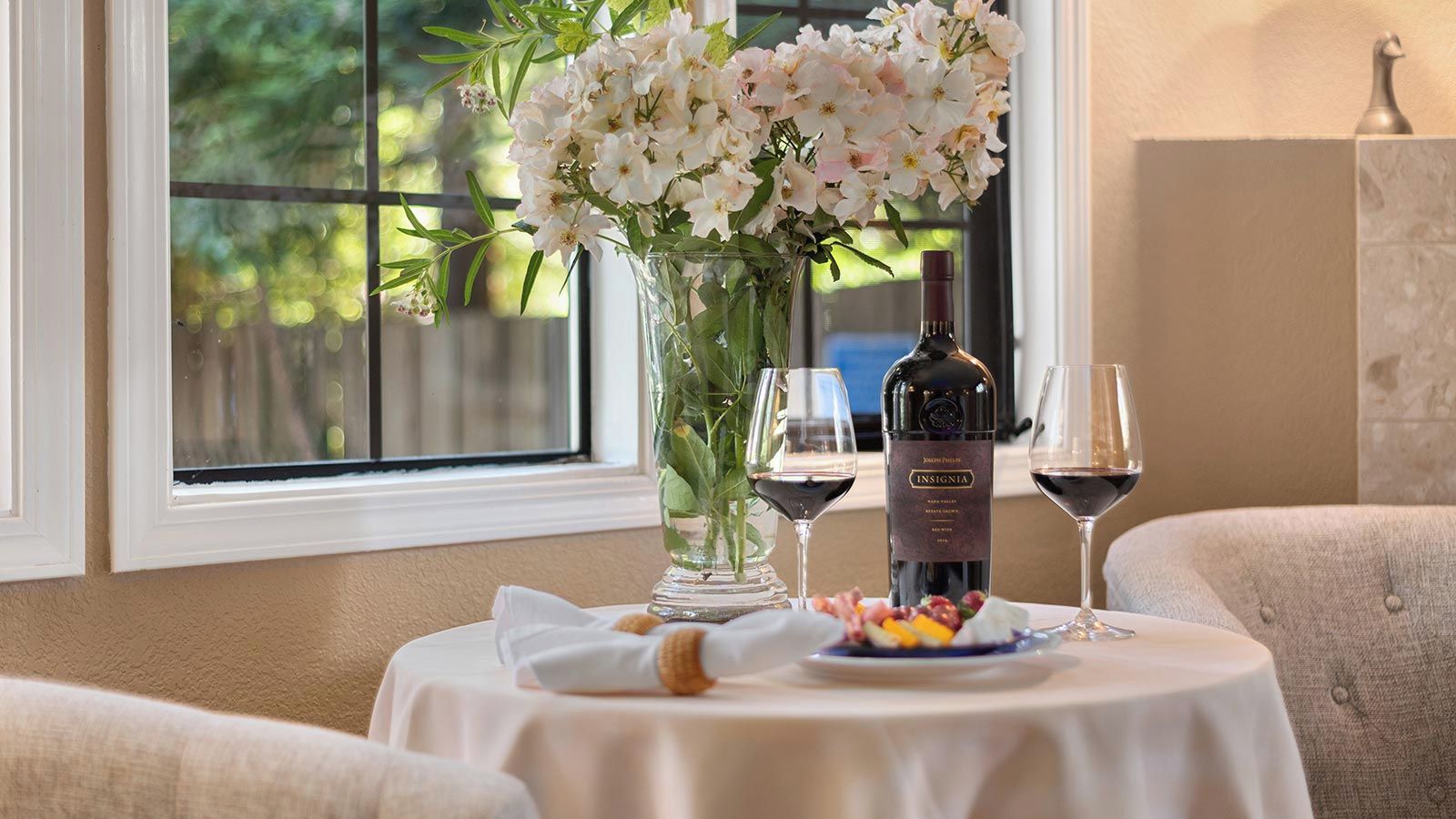 A dining table with a bottle of wine, glasses, a plate of food, and a vase of flowers.