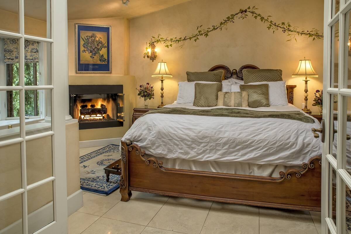 A bedroom with a large bed, two bedside lamps, a fireplace, and a framed floral artwork on the wall.