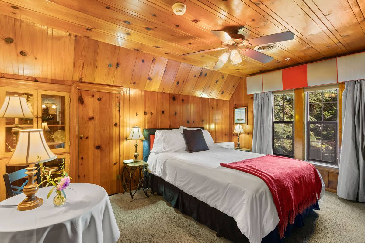 A wooden-paneled room with a bed, a round table with a lamp, and a window with curtains.