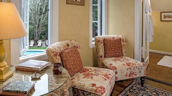 A room with two floral armchairs, a table with books and a decorative item, and a view of a pool outside. - Candlelight Inn Napa Valley (Napa)