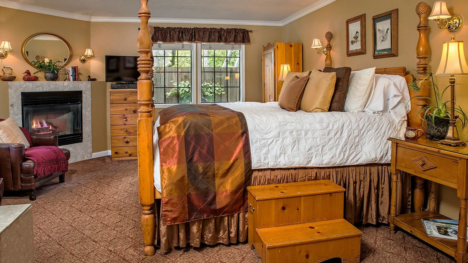 A bedroom with a four-poster bed, a fireplace, a television, and a window with a view of greenery.