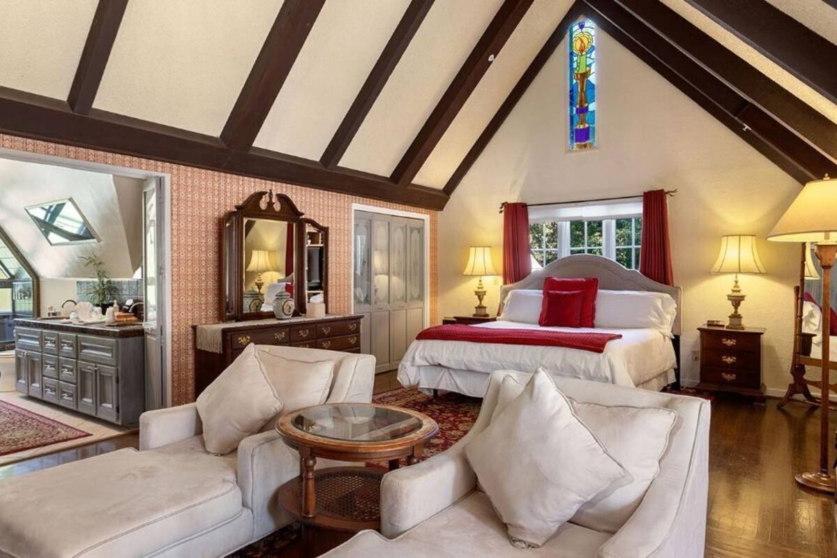A bedroom with a large bed, a sitting area with a glass table, a dresser, and a mirror.