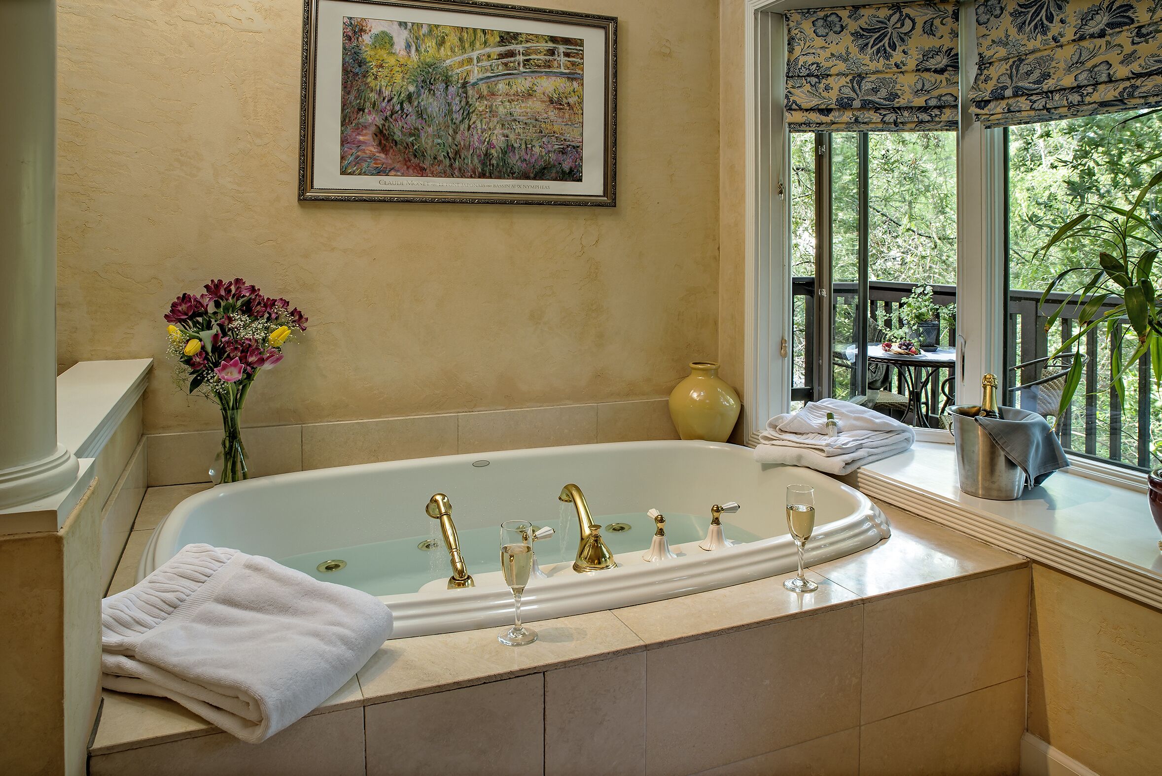 A bathroom with a large bathtub, a framed painting, and a window with a view of greenery.