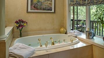 A bathroom with a large bathtub, a framed painting, and a window with a view of greenery.