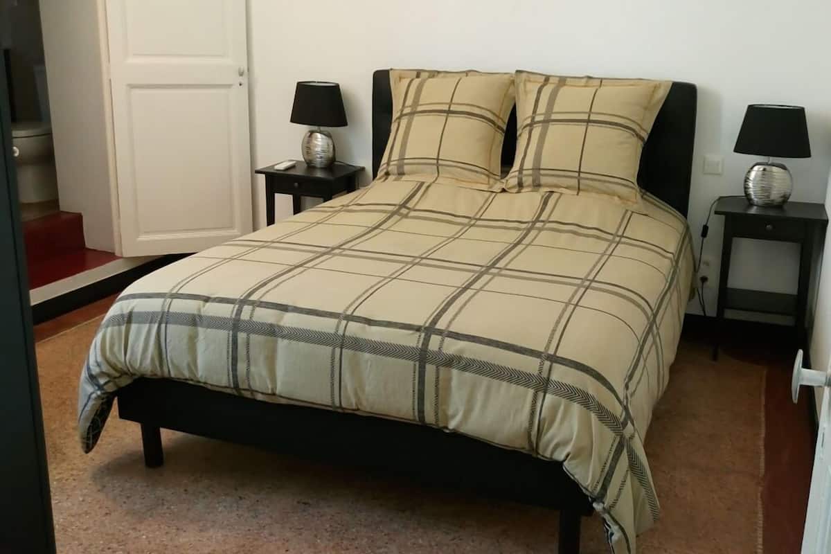 Uma cama bem arrumada com colcha xadrez, duas mesas de cabeceira com luminárias, e uma porta ao fundo.