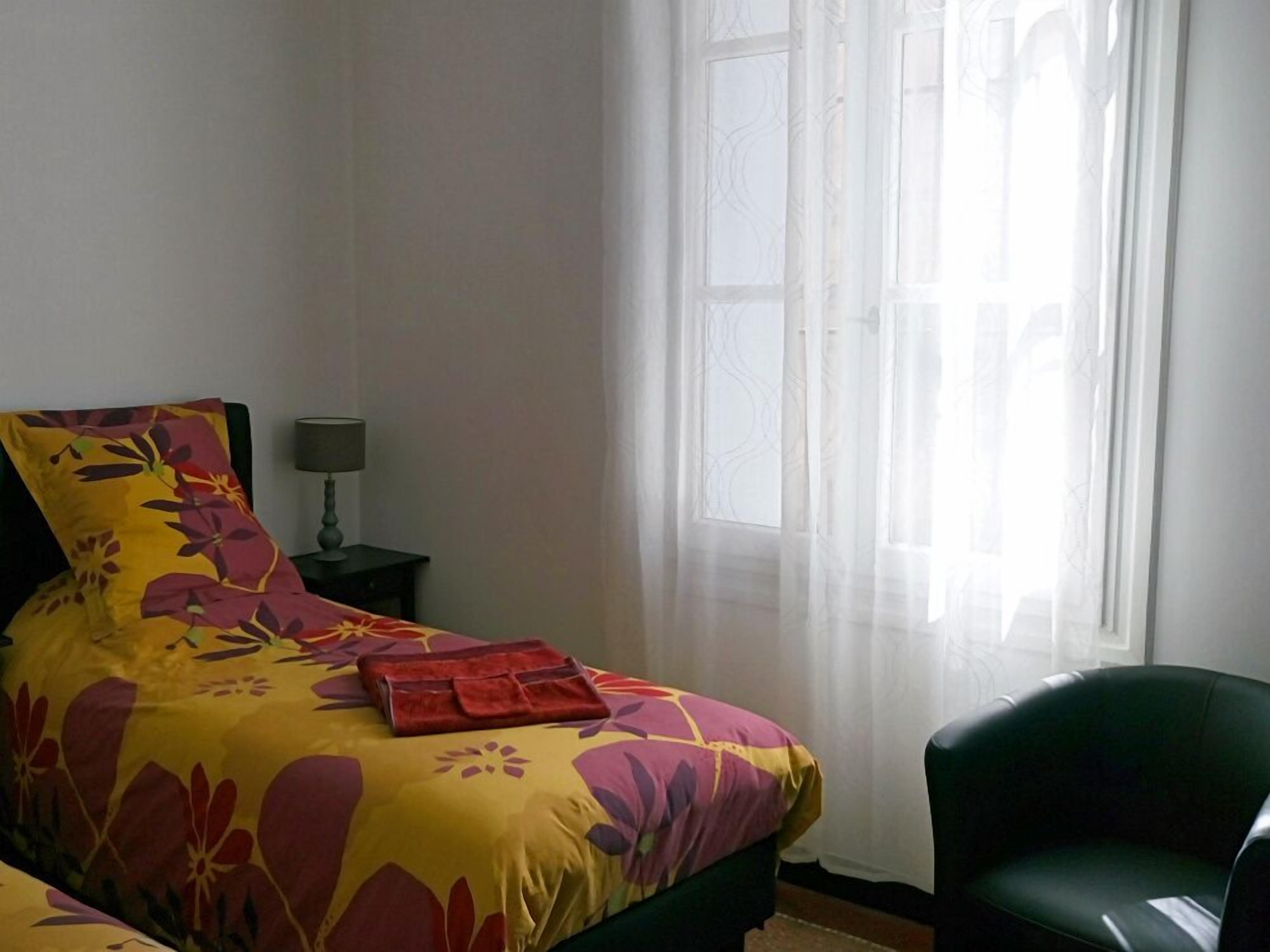 Une chambre à coucher avec un lit, une chaise, une table de chevet et une fenêtre ornée de rideaux légers.