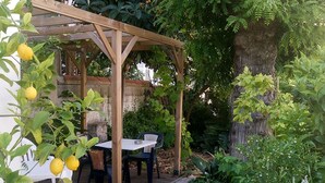 Une pergola en bois, avec une table et des chaises, entourée d’une végétation luxuriante et d’un tronc d’arbre.