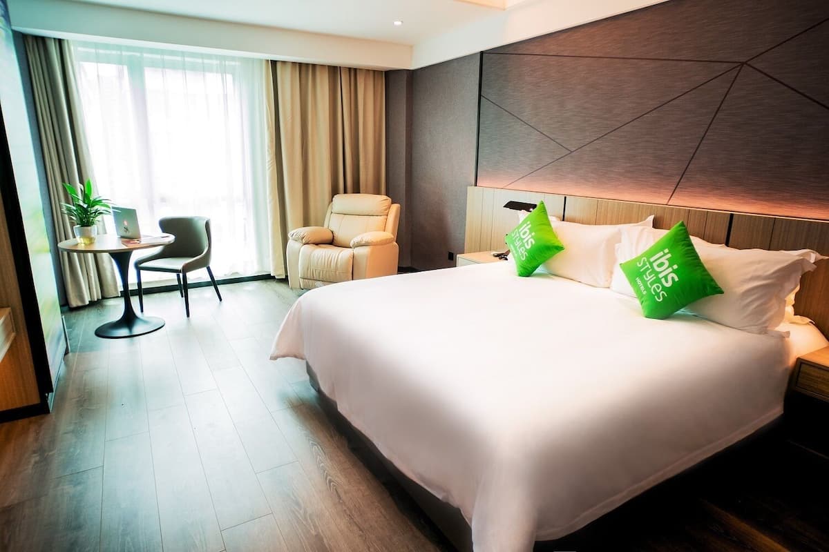 Ein Hotelzimmer mit einem großen Bett, zwei grünen Kissen, einem Nachttisch, einem Sessel, einem kleinen Tisch mit einer Pflanze und einem Fenster mit Vorhängen.